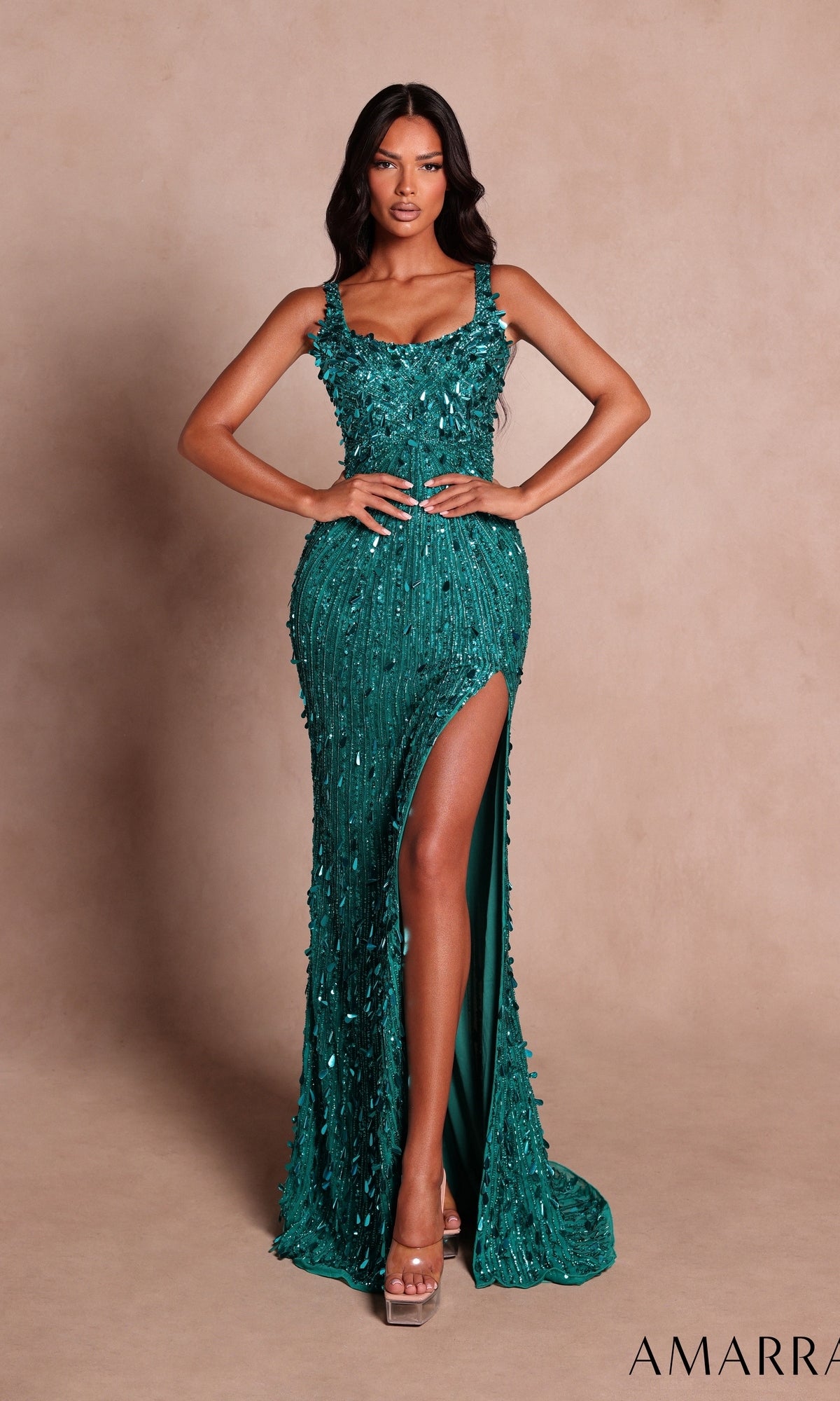 A woman wearing the Amarra 89775 long prom dress in jade—featuring a fitted silhouette, high slit, and textured beaded-style detailing—paired with clear high heels, stands against a plain beige background.